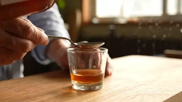 An older man's hand holding a tablespoon steady as dark amber liquid is poured into it